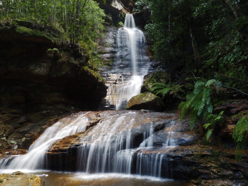 Empress Falls spilling out of Empress Canyon on the Valley of the Waters track in Wentworth Falls. Waterfall starts narrow at the top from the entrance to the canyon and widens as it spills down the rockface to a pool below, continuing to a form a smaller waterfall in front of the track. 
