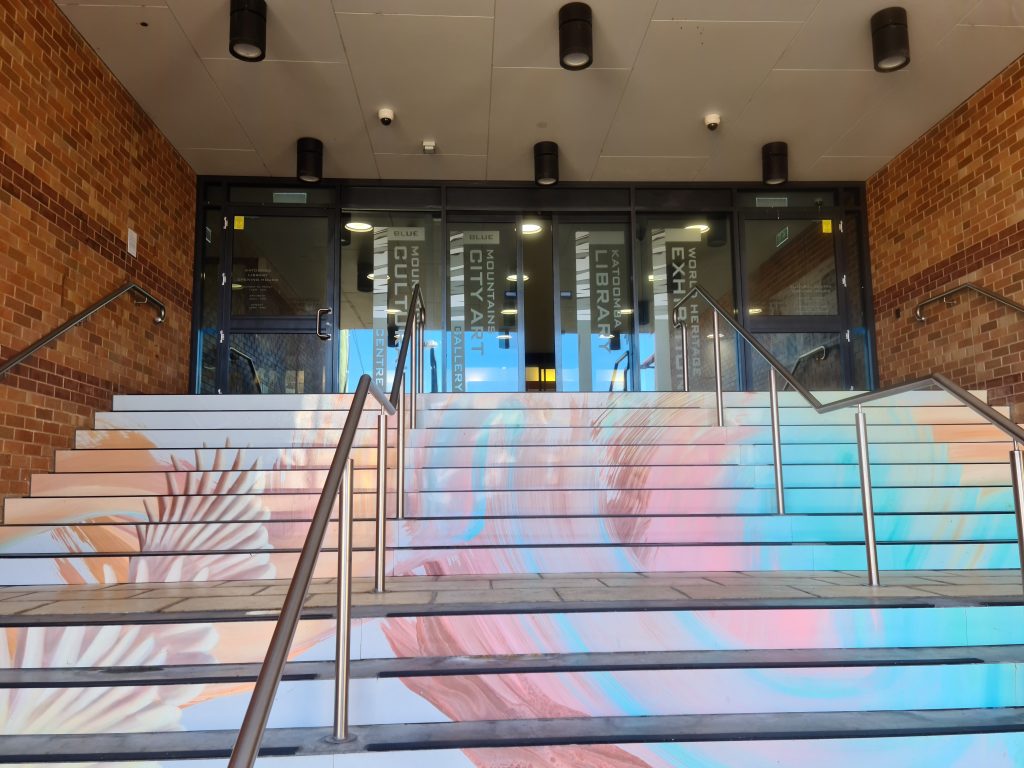 Pictured is the entry to the Blue Mountains Cultural Centre. Colour painted steps lead up to the glass sliding door entry. 