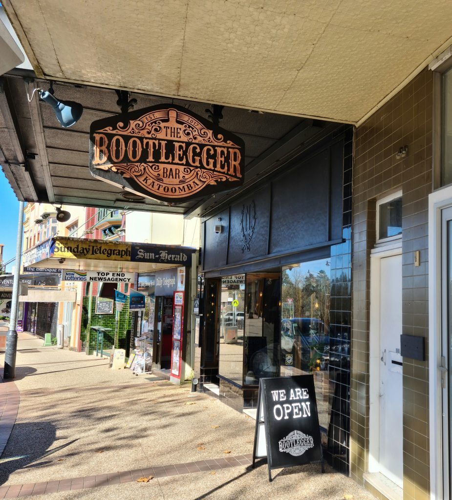 Pictured is the streetfront entrance of the Bootlegger Bar in Katoomba. A sign with Bootlegger in large writing is positioned above the street and the street frontage is all glass. 
