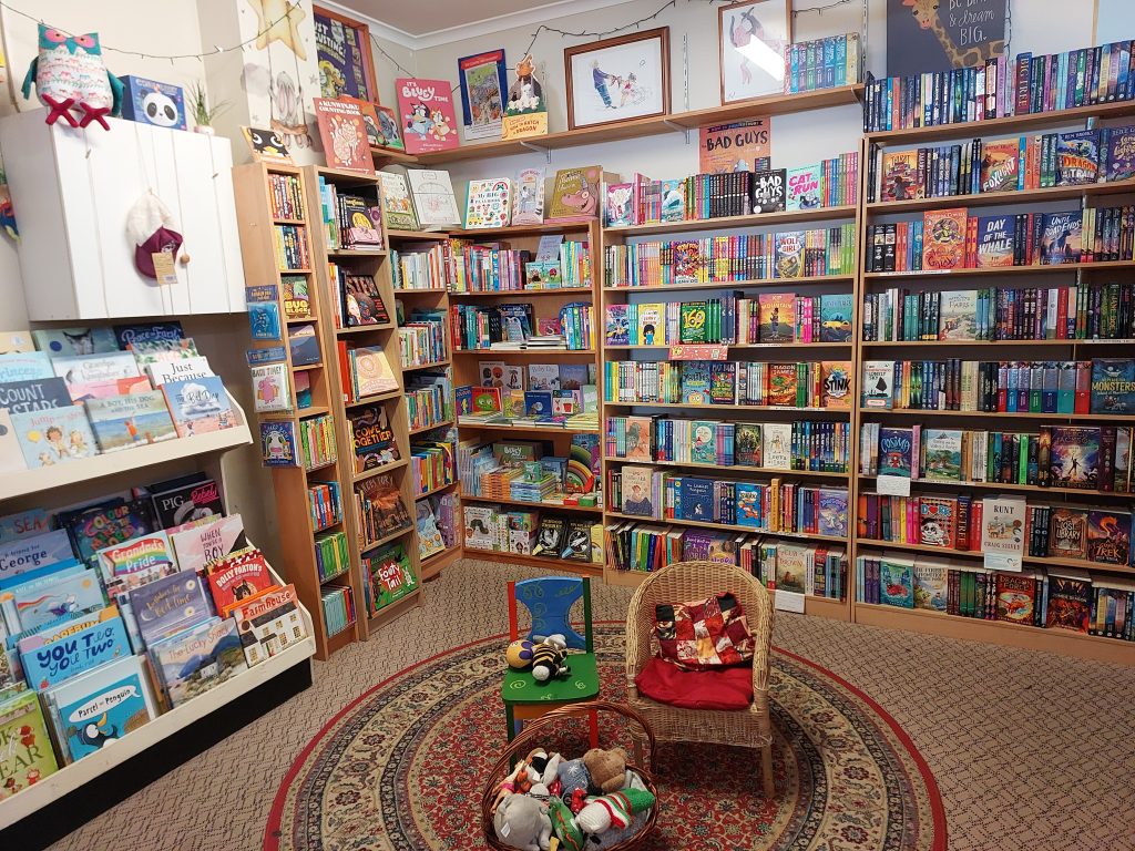 Image of a childrens corner in the Turning Page bookshop in Springwood. Two small chairs and a basket of toys are placed on a circular patterned rug and there are bookshelves behind.