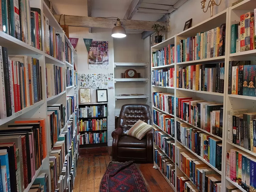 A corner in RoseyRavelston Bookshop. A maroon rug stretches across the centre of the floorboards and an armchair is at the end. Full bookshelves are on either side.