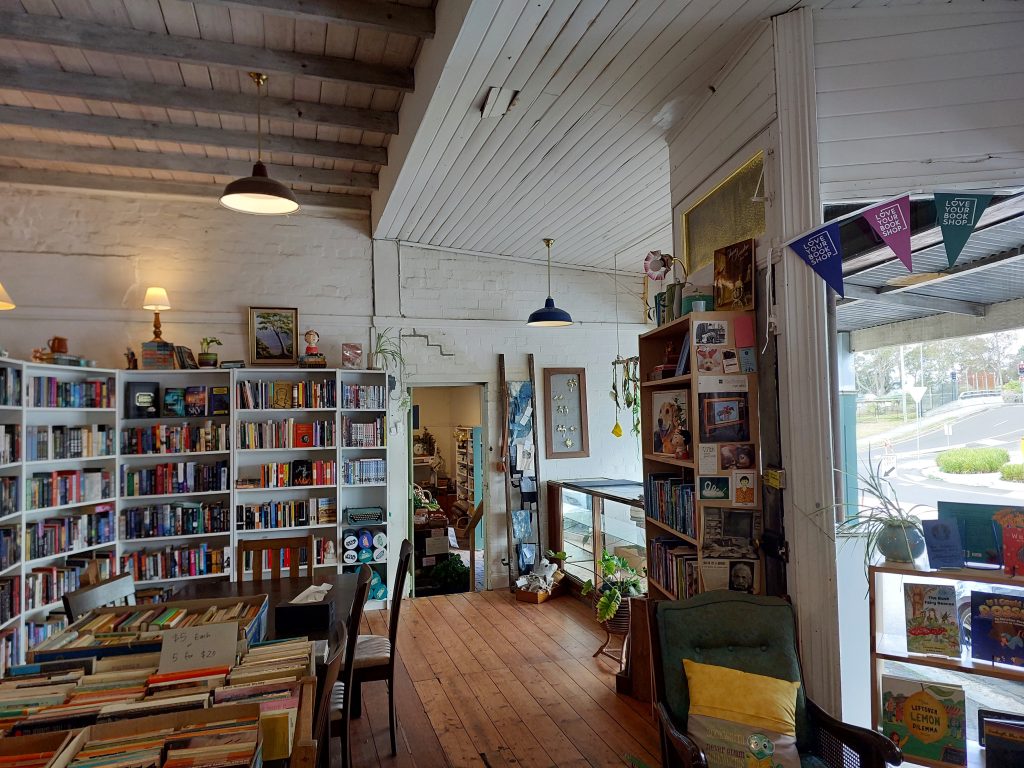 The main room in RoseyRavelston bookshop. A large table can be seen in the centre of the floor with natural light filling the room from the windows. Bookshelves stand full against the wall. 