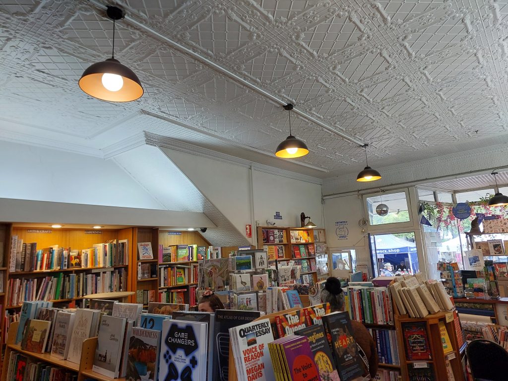 The interior of Megalong Books in Leura. Wooden shelves full of books can be seen, with a window looking out on Leura Mall. Lightshades with yellow white globes are hanging from the ceiling.